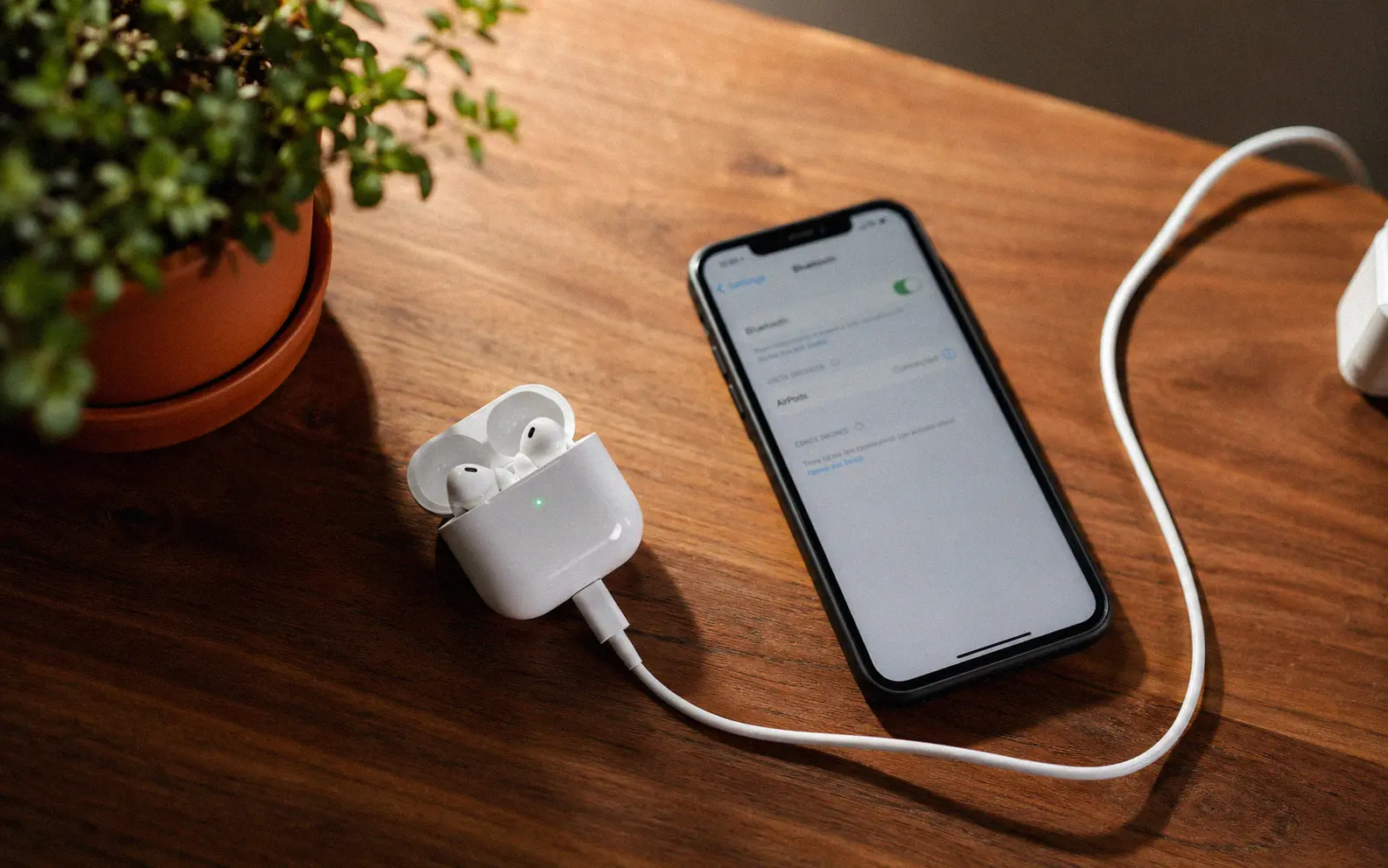 AirPods in open charging case connected to a Lightning cable on a wooden desk beside an iPhone showing Bluetooth firmware settings