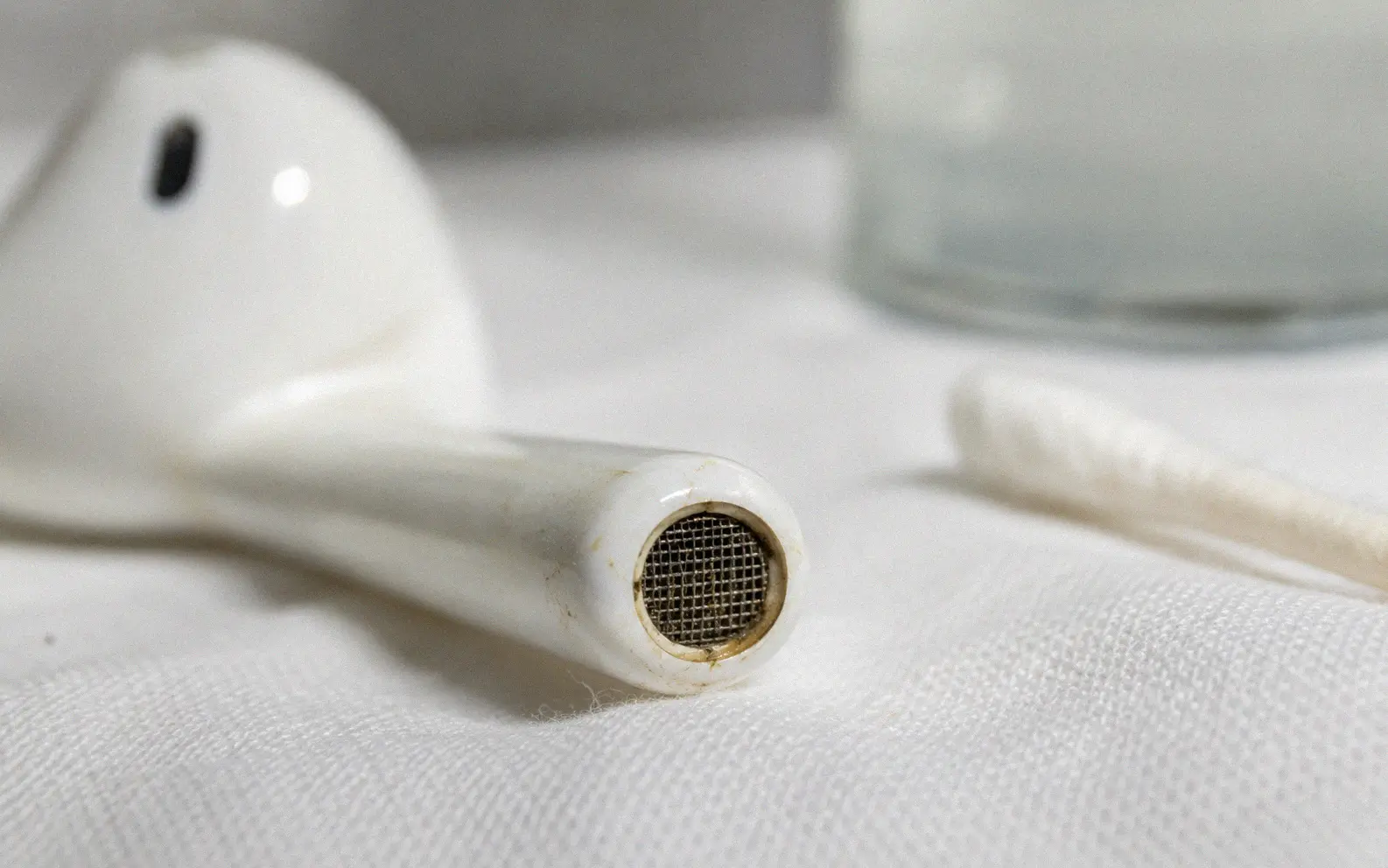 Close-up macro photograph of a white AirPod stem showing the microphone mesh grill with visible earwax debris and a cotton swab nearby