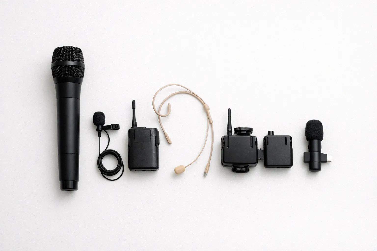 Five wireless microphone types laid out on a white desk including handheld lavalier headset camera-mount and phone clip-on